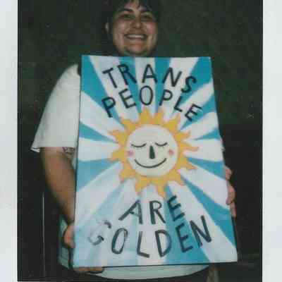 A person smiles, holding a colorful sign reading "Trans People Are Golden" with a sun illustration.