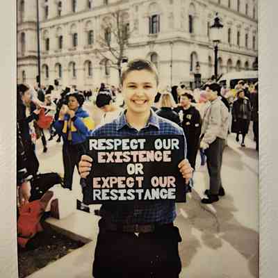 A person is holding a sign reading, "Respect our existence or expect our resistance," with a crowd behind them.