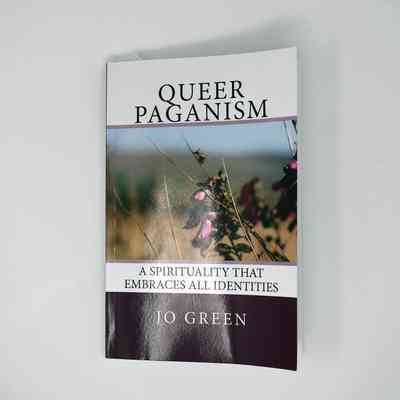 Front cover of a book titled "Queer Paganism - A spirituality that embraces all identities" by Jo Green. It also features a photo of a magenta flower and dry brush.