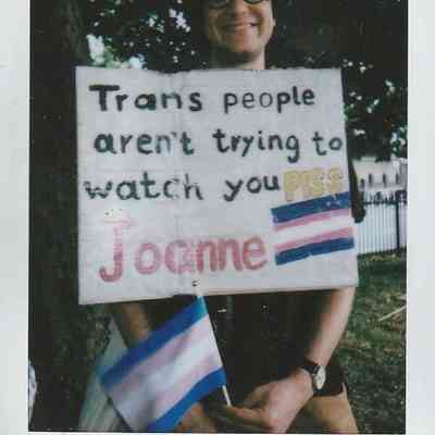 A person holds a sign which says "Trans People Aren't Trying To Watch You Piss Joanne" whilst they smile under a tree in a park setting.