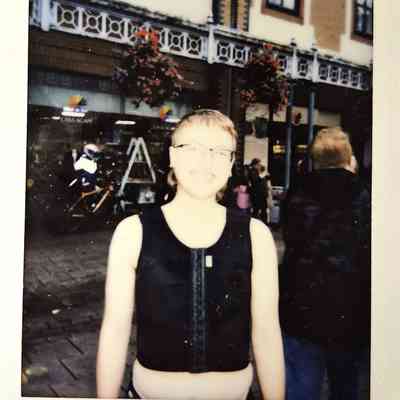 A person is smiling outdoors, wearing glasses and a black top, with buildings and people in the background.