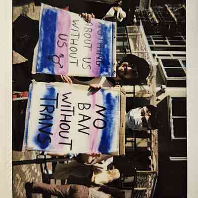 Two individuals hold signs which say "NOTHING ABOUT US WITHOUT US" and "NO BAN WITHOUT TRANS".