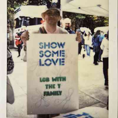 A person holds a sign saying “SHOW SOME LOVE LGB WITH THE T FAMILY” at an outdoor event.