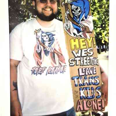 A person holds a colorful sign advocating for trans kids, wearing matching themed sunglasses and shirt.