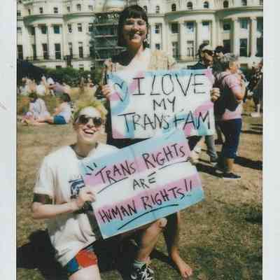 Two individuals in the sun smiling and holding two placards, one says "I Love My Trans Fam" and the other says "Trans Rights Are Human Rights".
