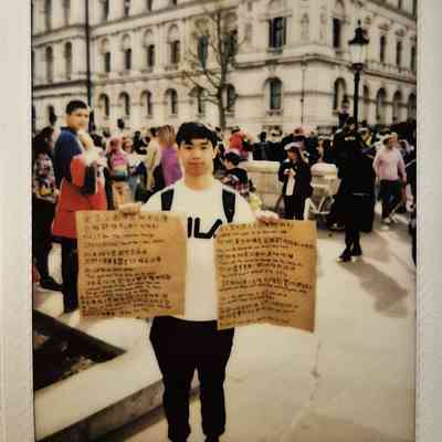 A person holds two written signs in a crowded public square, with a historic building behind them.