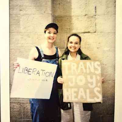 Two people are holding signs saying “LIBERATION” and “TRANS JOY HEALS”, smiling against a wall.