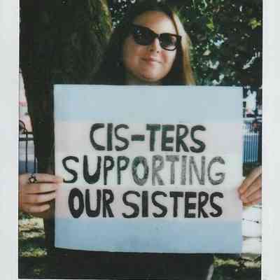 A person wearing sunglasses holds a sign that reads "Cis-ters Supporting Our Sisters," standing outdoors