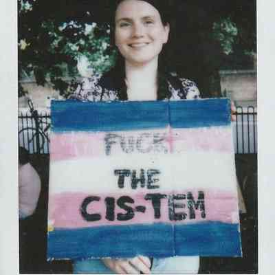 A person holds a colorful protest sign with a message challenging societal norms in a park setting.