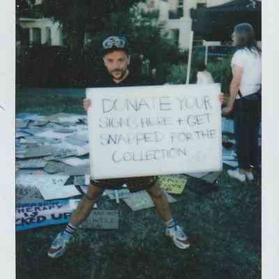 E-J Scott holds a placard which says "DONATE YOUR SIGNS HERE + GET SNAPPED FOR THE COLLECTION". Behind E-J is a grass field full of placard on the floor.