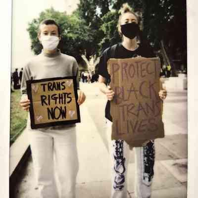 Two individuals, wearing masks and holding protest signs, stand in support of trans rights and equality.