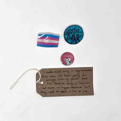 The image shows badges and a handwritten tag describing a personal transition journey.
