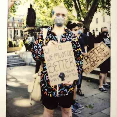 A person stands in a colorful hoodie, holding a protest sign amidst a group of people in a park