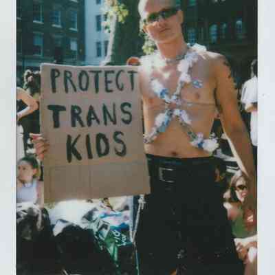 A person stands outdoors holding a cardboard sign that reads "Protect Trans Kids" during a sunny day.