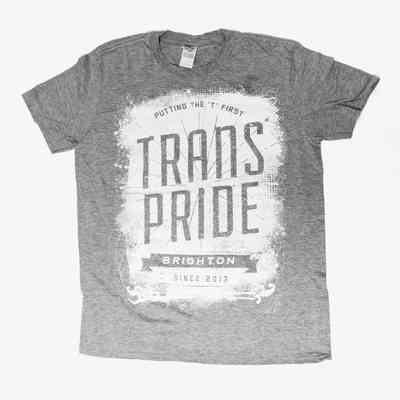 A grey T-shirt with the text "Putting the 'T' First" at the top, "Trans Pride" in large letters in the middle "Brighton" on a banner underneath, and "Since 2013" at the bottom. A label with the brand "Gildan" can be seen on the neck.