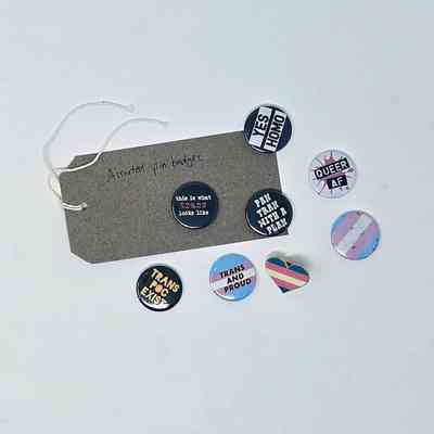 Assorted pin badges with various pride slogans and designs are displayed on a white surface.