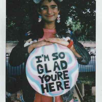 A person with colorful accessories holds a sign reading, "I'M SO GLAD YOU'RE HERE".