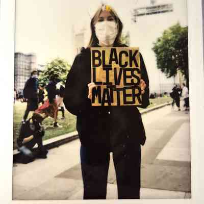 A masked individual holds a "Black Lives Matter" sign during a public demonstration outdoors with others nearby.