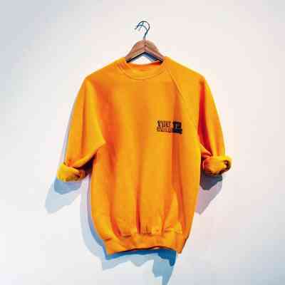 A yellow sweatshirt with rolled up sleeves and "THE TIN WARDROBE" printed on the breast, on a wooden hanger.