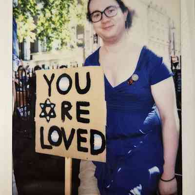 A person in a blue dress holds a sign reading "YOU ARE LOVED" at a protest.