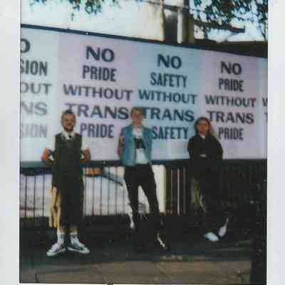 Three people stand in front of signs which say "No Pride Without Trans Pride".