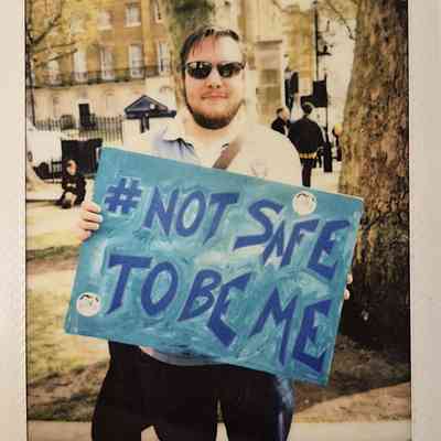 A person holding a sign that reads "#NOT SAFE TO BE ME" stands outside, wearing sunglasses.