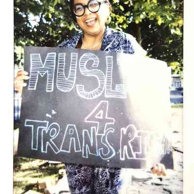 A smiling individual holds a sign that reads, "MUSLIMS 4 TRANS RIGHTS,"
