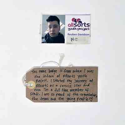 Name badge & tag; Allsorts Youth Project; name "Reuben Davidson" with photo wearing black scarf. Sticker handwritten "He". Card laminated, repaired in sellotape on back. Card creased across face in photo.