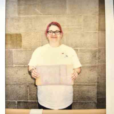 A person with pink hair and glasses holds a sign, standing against a textured stone wall background.