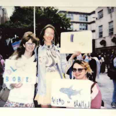 Three people are smiling and holding signs in a lively outdoor setting at a public event.