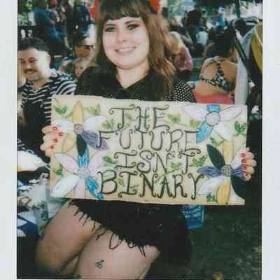 A person holds a sign saying "THE FUTURE ISN'T BINARY" surrounded by colorful flowers.