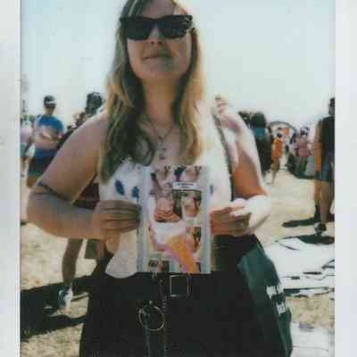 A woman wearing sunglasses holds a colorful magazine, standing outdoors among a crowd on a sunny day.