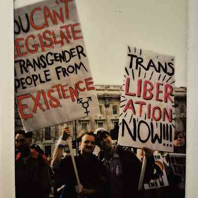 The image shows people holding signs advocating for transgender rights and liberation including one sign which says "TRANS LIBERATION NOW!!"