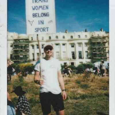 A person is holding a sign stating "Trans Women Belong in Sport" in an outdoor setting.
