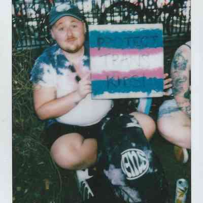 A person sits on the ground, holding a sign that reads "Protect Trans Kids" with colorful background