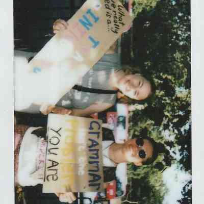 Two individuals hold signs with messages at an outdoor event, smiling and surrounded by trees.
