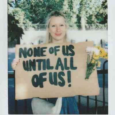 A person holds a cardboard sign reading "None of us until all of us!" with flowers attached.