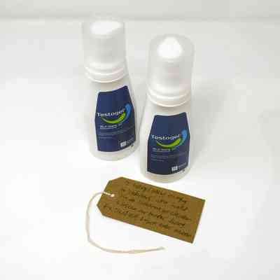 Two bottles of Testogel. The bottles are white and have a dark blue label with a green and light blue splash logo. With a brown, handwritten luggage tag.