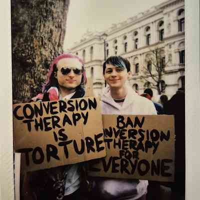 Two people hold protest signs which says "CONVERSION THERAPY IS TORTURE" and "BAN CONVERSION THERAPY FOR EVERYONE".