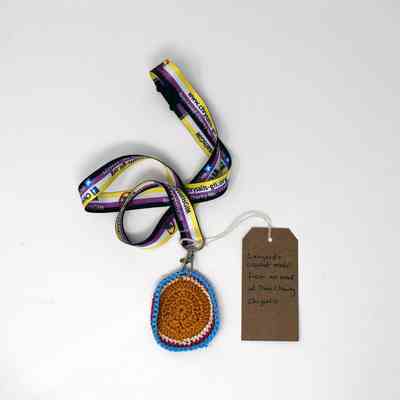 A colorful lanyard with a crocheted medal and a brown tag on a white background.