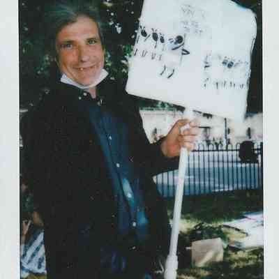 A person stands in a park holding a sign with illustrations depicting trans people.