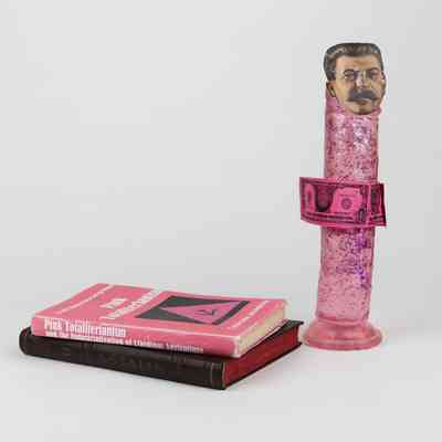 A pink, glitter-covered dildo with a a cutout of Stalin's face on the top stands beside two books stacked on top of each other. The top book is pink with the title "Pink Totaliterianism."