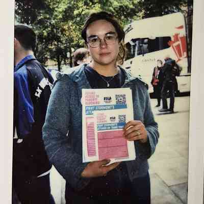 A person holding flyers stands outside, with trees and a bus in the background while wearing glasses.