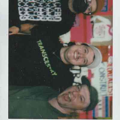 Three people are happily posing together, wearing casual clothes and hats, against a colorful background.