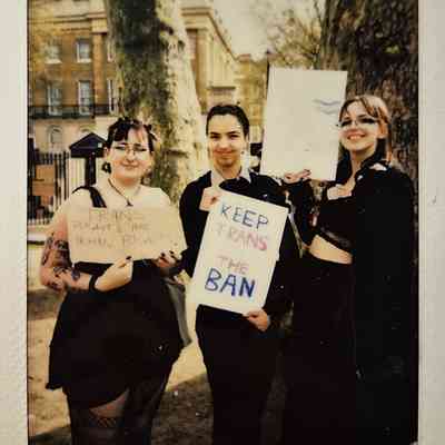 Three people are holding signs advocating for trans rights with phrases such as "KEEP TRANS IN THE BAN" beside large trees outdoors.