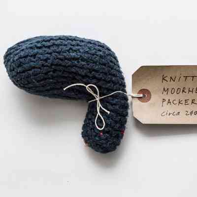 Dark blue knitted packer. a few bits of orange yarn poke through the blue stitches at the top tight of the packer. Thers is a white tag tied around the top.