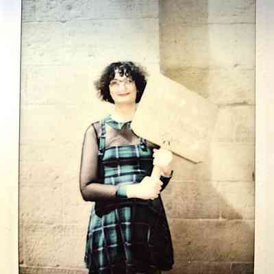 A person stands holding a sign, wearing glasses and a plaid dress, against a stone wall.