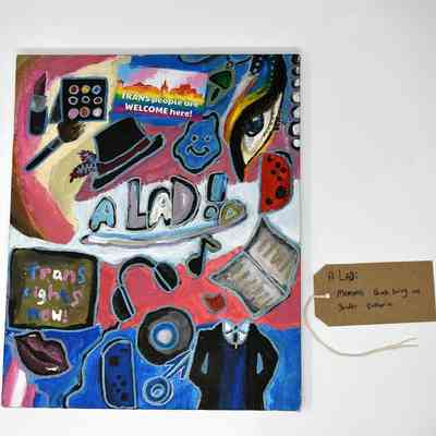 The colorful artwork features symbols and messages supporting transgender rights with the text "A LAD!" in the centre. With a brown handwritten luggage tag.