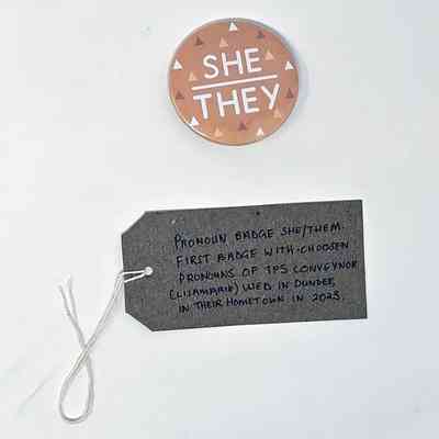 The image shows a button with "She/They" above a gray tag with handwritten notes.