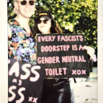 Two people are holding signs with a message about fascists, in a sunlit outdoor setting.